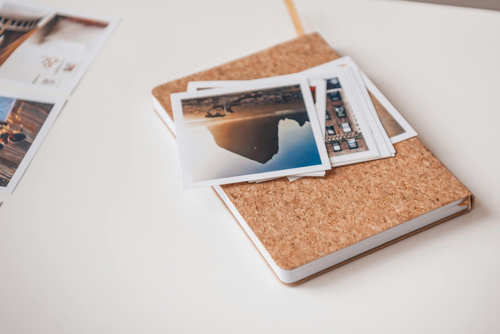 Cork-covered notebook topped with printed photographs on a white desk, conveying travel memories.